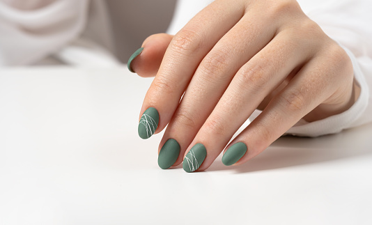 Hands of a young woman with green olive matte nails on a light gray background. Manicure, pedicure beauty salon concept. Copy space for text or logo. Gel polish and abstract white spider web pattern.