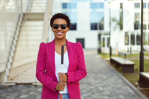 Portrait of a confident and cheerfulmixed-race businesswoman, with visual impairment, standing in front of her corporate building while holding blind person cane