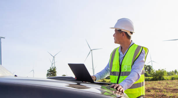 istock front view Asian engineer smiling holding folder looking at camera with windmill, wind turbine power background. 1323184666