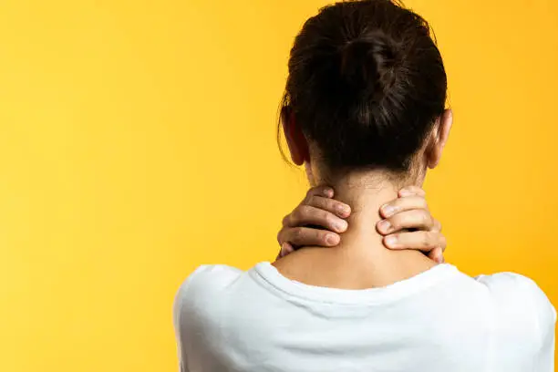 Back view of unrecognizable woman with neck pain in front of yellow background.