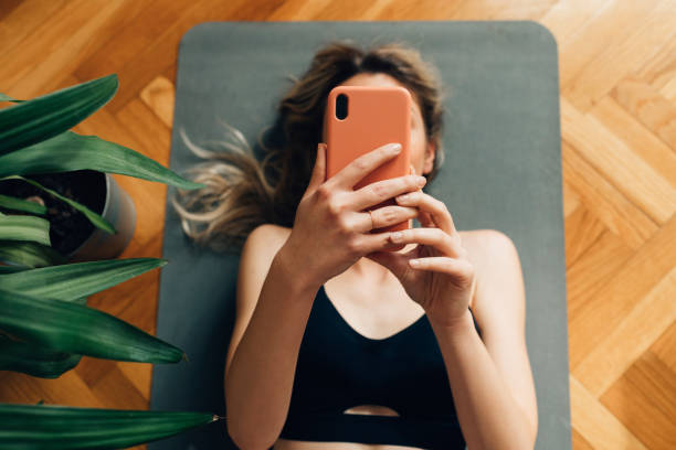 A young Caucasian woman relaxing after workout on an exercise mat and checking social media on her mobile phone