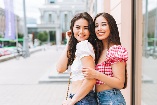 Happy smiling beautiful brunette young women friends in summer clothes walking on the summer city street