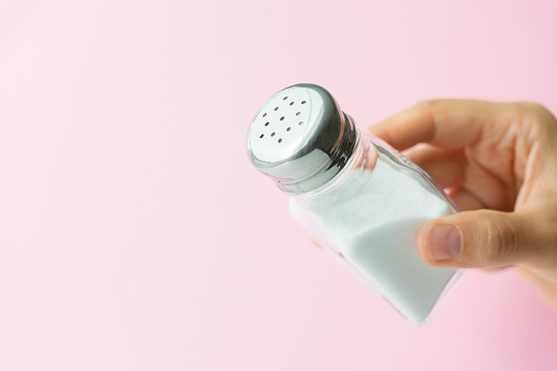 Hand is holding salt shaker with salt on soft pink background.
