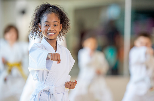 A young African American girl practices karate in a class setting and smiles at the camera.