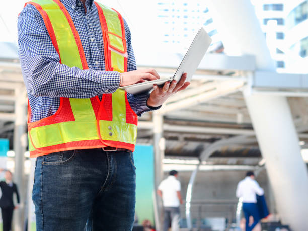 istock Young Asian male engineer wearing safety vest and helmet, standing and using laptop computer at modern city, young staff man worker working at the street outside. 1323209336