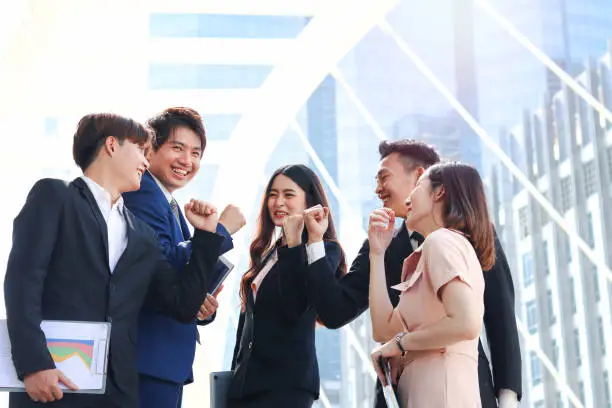 Group of five young Asian cheerful business people raising fist hands up for celebrating success, businessman and woman cheer up their colleagues as commitment of strong teamwork at outside with skyscraper building as blurred background.