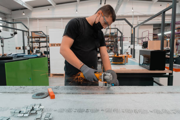 istock Heavy Industry Engineering Factory Interior with Industrial Worker Using Angle Grinder and Cutting a Metal Tube.He Wears a Mask on His Face Because of the Coronavirus Pandemic 1323214254
