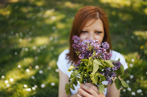 Happy young woman in white T-shirt standing in public park, smiling and smells lilac