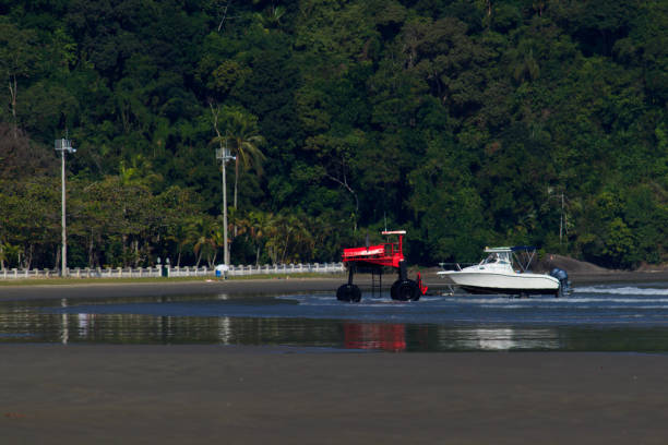 in the foreground, a strip of wet sand reflecting a boat with a white hull towed by a red tractor, close to a hill covered with native vegetation from the atlantic forest. - sao paulo south america marina southeastern region imagens e fotografias de stock