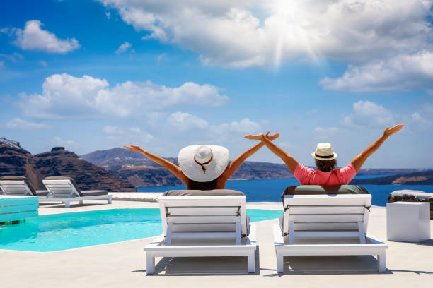 Happy vacation couple sitting on white sunchairs by the pool Happy vacation couple sitting on white sunchairs by the pool and enjoying the sun during their summer holidays in Santorini, Greece happy couple on vacation in santorini greece stock pictures, royalty-free photos & images