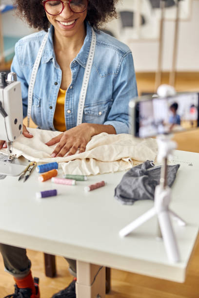istock African-American lady designer sews with machine shooting video at table in studio 1323232777