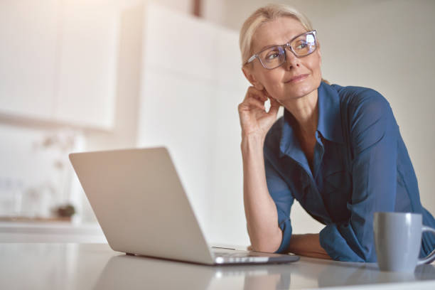 istock Pensive mature businesswoman looking aside, working from home using laptop computer and drinking coffee in the morning, standing in the kitchen 1323233967