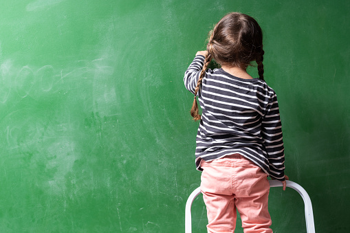 Rear photo of preschooler girl wearing a striped sweater writing on green chalkboard. The blackboard is blank. Shot indoor with a full frame mirrorless camera.