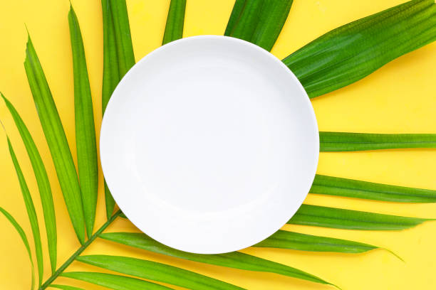istock Empty white ceramic plate on tropical palm leaves on yellow background. 1323240417