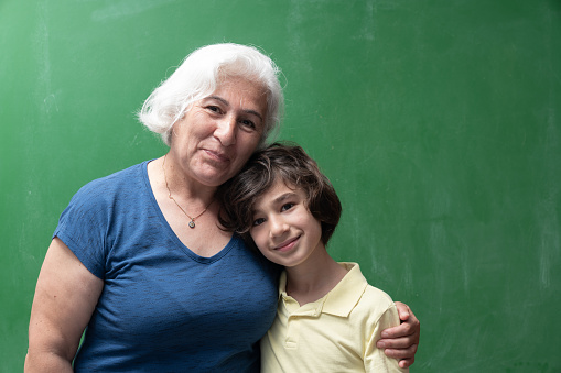 Portrait of senior grandmother with white hair hugging her 8 years old grandson in front of green chalkboard.. The board şs blank. Shot with a full frame mirrorless camera.