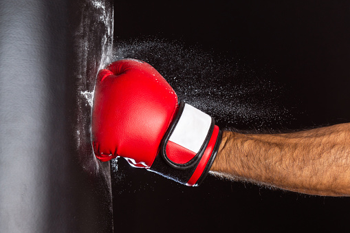 fist of a man with red boxing glove hitting a black punching bag on black background