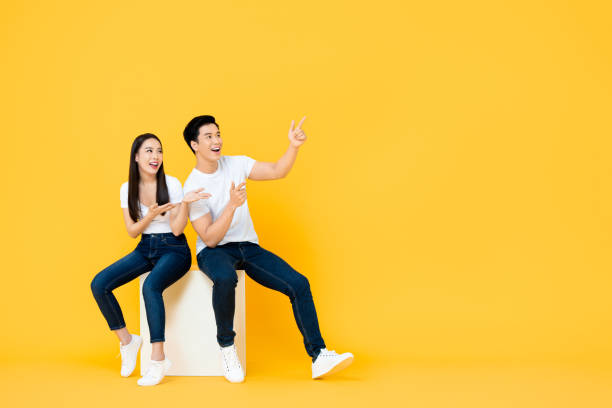 Happy surprised attractive young Asian couple pointing and looking at blank space beside in yellow isolated stuidio background Happy surprised attractive young Asian couple pointing and looking at blank space beside in yellow isolated stuidio background girl sitting stock pictures, royalty-free photos & images