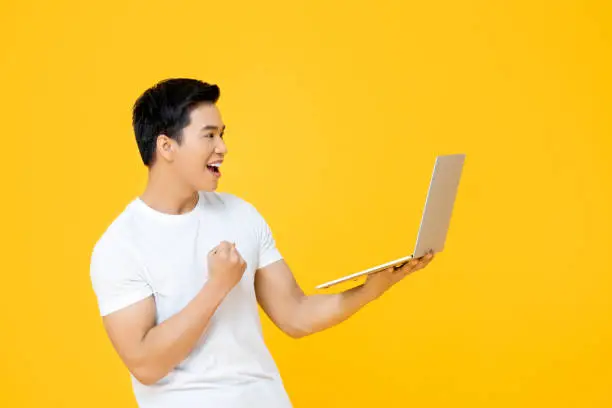Happy young Asian man looking at laptop computer and raising his fist doing yes gesture isolated on yellow background