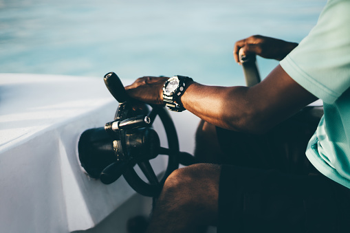A close-up view of a hand with the watch of a black adult guy turning the steering wheel while driving a motorboat on a sunny evening, selective focus, shallow depth of field