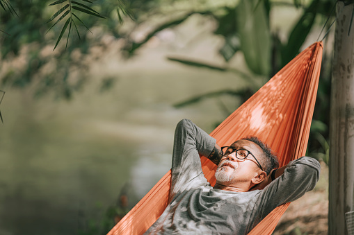 asian chinese senior man enjoying his afternoon lying down at hammock at riverside under bamboo trees