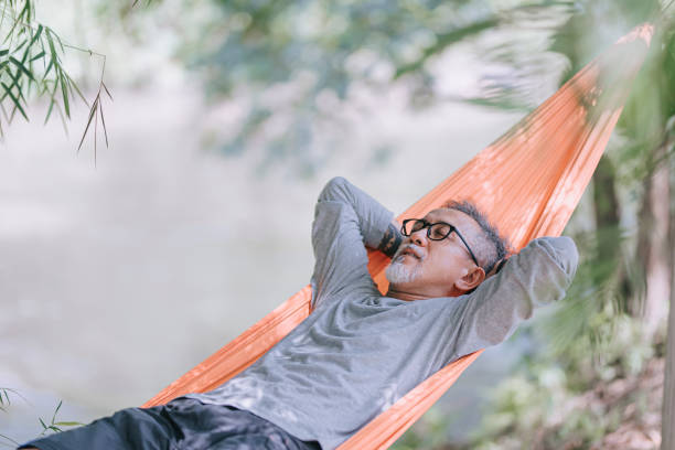 asiatische chinesische senior mann genießt seinen nachmittag liegend an hängematte am flussufer unter bambusbäumen - hängematte stock-fotos und bilder