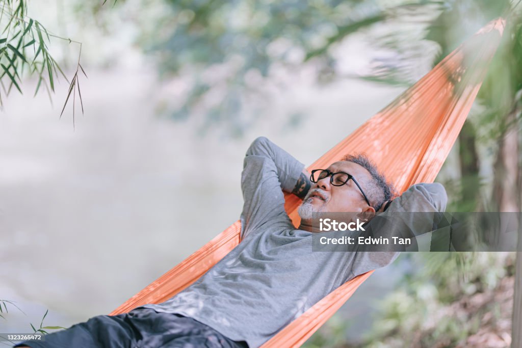asian chinese senior man enjoying his afternoon lying down at hammock at riverside under bamboo trees Relaxation Stock Photo