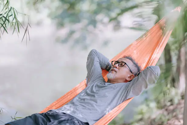 Photo of asian chinese senior man enjoying his afternoon lying down at hammock at riverside under bamboo trees