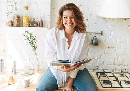 Gorgeous happy young woman plus size body positive in blue jeans and white shirt reading cooking book in home kitchen