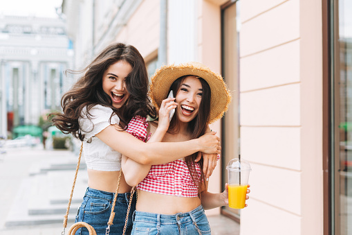 Happy smiling beautiful brunette young women friends in summer clothes with juice in hands having fun on the summer city street