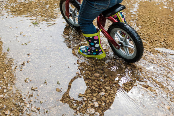 istock Child playing on water puddle with a toy bicycle and spot rain boots 1323285673