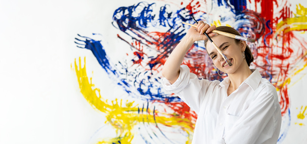 Painting course. Art banner. Fun hobby. Happy playful smiling artist girl in white with paintbrushes on colorful yellow blue red abstract messy wall background with copy space.