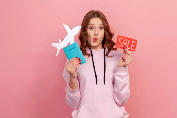 Portrait of excited curly haired teenage girl in hoodie showing passport with paper airplane and sale word sign, buying cheap tickets, discount. Indoor studio shot isolated on pink background