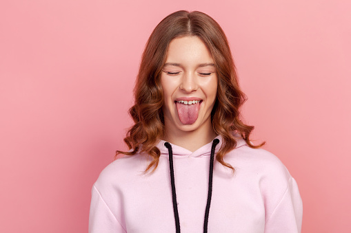 Portrait of adorable teenage girl in hoodie with eyes closed showing tongue, looking playful naughty, female millenial having fun. Indoor studio shot, isolated on pink background