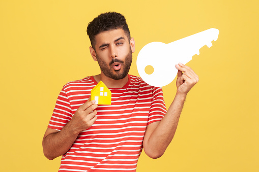 Happy satisfied man home owner with beard in striped t-shirt winking holding paper house and key, estate purchase or mortgage. Indoor studio shot isolated on yellow background