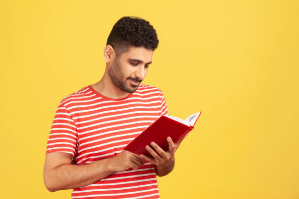 istock Concentrated bearded man holding book and smiling, enjoying reading, standing absorbed with exciting plot, checking schedule in notepad. 1323294795