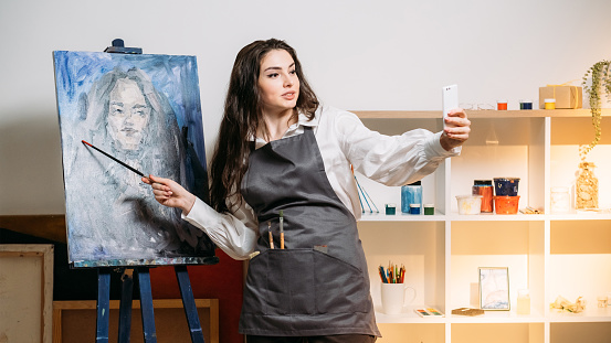 Art studio. Female artist. Home painting. Creative profession. Beautiful woman pointing brush on canvas portrait on easel making selfie smartphone light room interior.