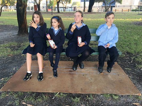 Primary school children sitting on a bench  eating food during recess.