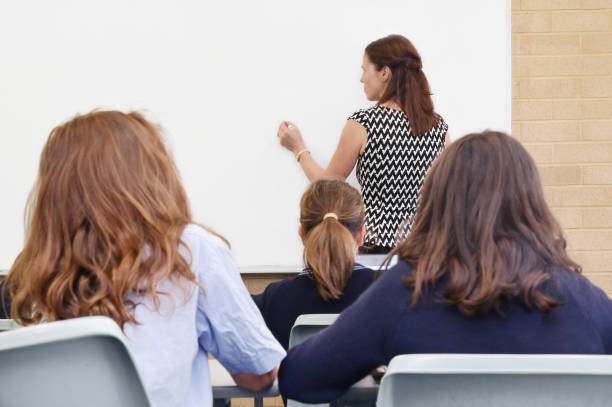 Primary school teacher writing on a clear whiteboard in classroom Rear view of primary school teacher writing on a clear whiteboard in classroom with students. only girls stock pictures, royalty-free photos & images