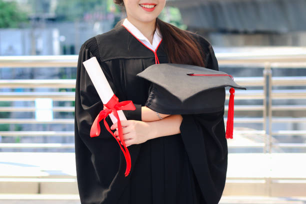 Happy smiling graduated student, young beautiful Asian woman holding square academic hat cap and certificate, standing with arms crossed at city with skyscraper building background, celebrating successful education on commencement graduation day when finished studies for university or college. feliz estudiante graduada sonriente, joven hermosa mujer asiática sosteniendo gorra de sombrero académico cuadrado y certificado, de pie con los brazos cruzados en la ciudad con el fondo de construcción de rascacielos, celebrando la educación exitosa e