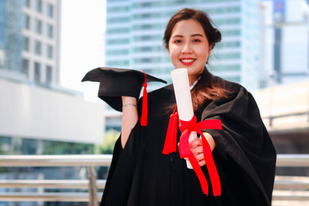 Happy smiling graduated student, young beautiful Asian woman holding square academic hat cap and giving certificate to camera, standing at city with skyscraper building background, celebrating successful education on commencement graduation day when finished studies for university or college. feliz estudiante graduada sonriente, joven hermosa mujer asiática sosteniendo gorra de sombrero académico cuadrado y dando certificado a la cámara, de pie en la ciudad con fondo de construcción de rascacielos, celebrando la educación exitosa en el dí