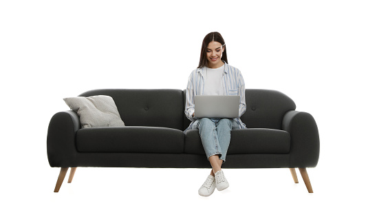 Young woman with laptop on comfortable grey sofa against white background