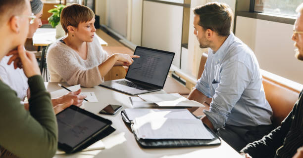 Female executive showing data to team in meeting Manager pointing at laptop screen and talking to colleagues during business meeting. Female executive showing data to team in meeting. finance stock pictures, royalty-free photos & images