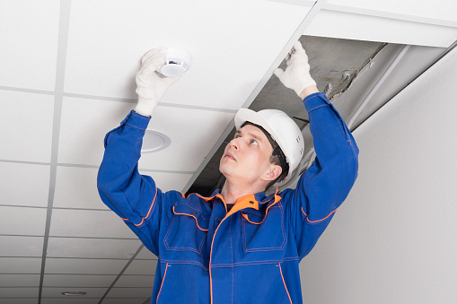 a worker installs a smoke and fire detector on the ceiling in the office