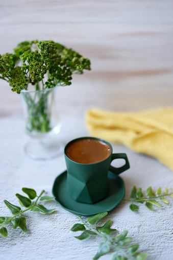 Istanbul, Turkey-June 12, 2021: Foamy Turkish coffee in a dark green cup on a white rough concrete floor. Small green leaves around and a yellow linen napkin. There are green plants in the vase. Shot with Canon EOS R5.