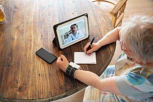 Senior woman sitting at wooden table in living room and talking with doctor from home via online video call about results of blood pressure measurement