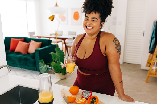 Happy female holding glass of fresh fruit juice while standing at kitchen. Woman in sports clothing having fruit juice at home.