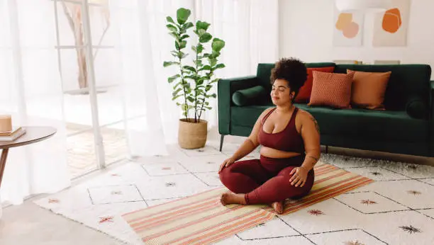 Body positive woman sitting cross legged and closed eyes in living room. Female in sports clothing practicing meditation at home.