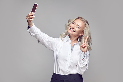 fashionable studio portrait of pretty and cheerful senior blonde woman wearing in stylish casual clothing of classic strict style, taking self portrait on phone over gray isolated background.