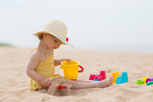 Little girl in yellow swimsuit and straw hat sitting on sand and playing with bucket and spade at sea beach in warm sunny summer day. Close up. Side view.