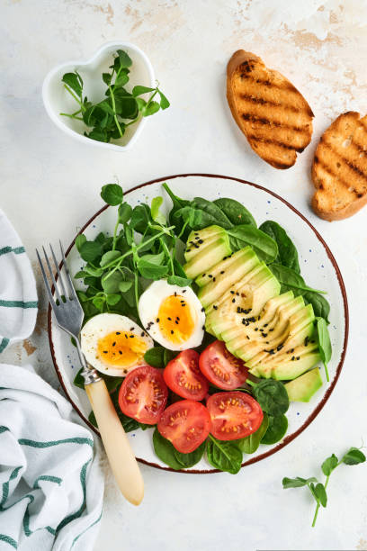 istock Avocado, cherry tomato, spinach and chicken egg, microgreens peas and black sesame seeds fresh salad in bowl on white stone table background. Healthy breakfast food concept. Top view. 1323369088